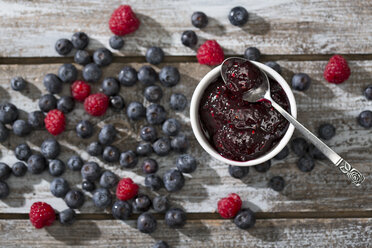 Schale mit Blaubeer- und Himbeermarmelade, Löffel, Himbeeren und Blaubeeren auf Holztisch, Blick von oben - MAEF008009