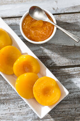 Bowl of peach jam and plate with halves of peaches on wooden table, elevated view - MAEF008041