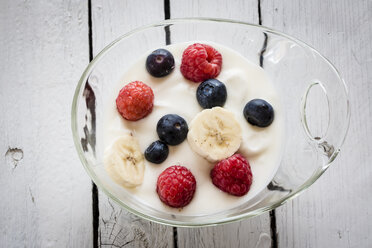 Glass bowl of natural yoghurt with blueberries, raspberries and banana slices on wooden table - SARF000277