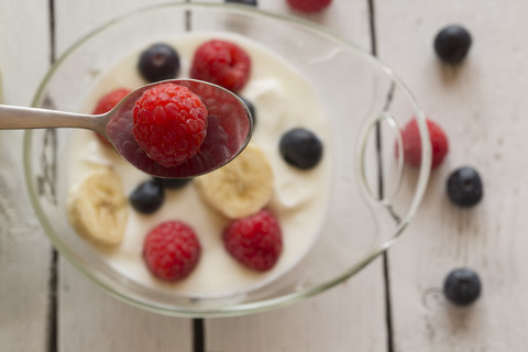 Löffel mit einzelner Himbeere vorne und Glasschale mit Naturjoghurt mit Heidelbeeren, Himbeeren und Bananenscheiben auf Holztisch, lizenzfreies Stockfoto