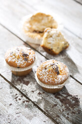 Zwei Muffins in Pappbechern und ein halber Muffin im Hintergrund auf grauem Holztisch - MAEF008049