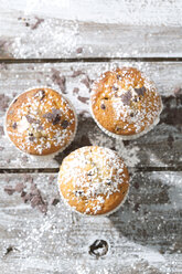 Drei Muffins in Pappbechern, bestreut mit Puderzucker und Schokoladenspänen auf einem Holztisch, Blick von oben - MAEF008063