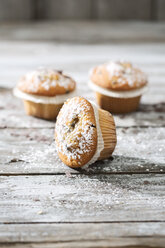 Drei Muffins in Papierförmchen mit Puderzucker bestreut auf Holztisch - MAEF008062