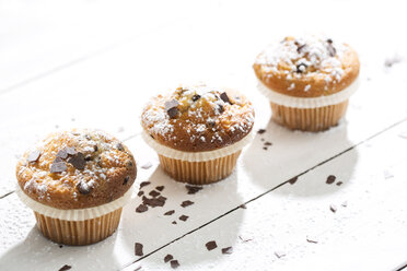 Drei Muffins in Pappbechern, bestreut mit Puderzucker und Schokoladenspänen auf weißem Holztisch - MAEF008059