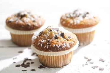 Drei Muffins in Pappbechern, bestreut mit Puderzucker und Schokoladenspänen auf weißem Holztisch - MAEF008058