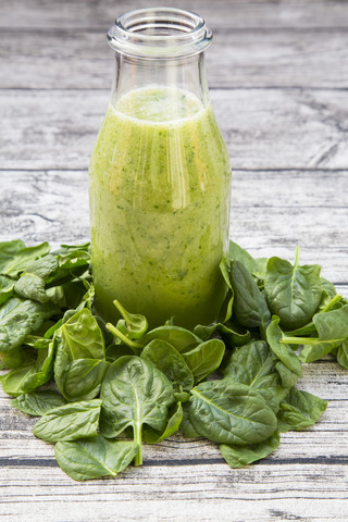 Glasflasche mit grünem Smoothie aus Spinat, Rucola, Apfel, Orange, Banane und Gurke inmitten von Babyspinatblättern auf grauem Holztisch, lizenzfreies Stockfoto