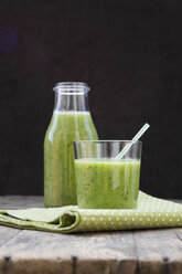 Glass bottle and glass of green smoothie, made of spinach, rocket salad, apple, orange, banana and cucumber, on wooden table - LV000781