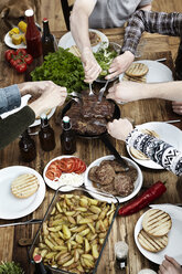 Friends eating potatoes, steaks and meatballs at wooden table - FMKF001074