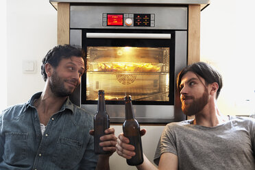 Zwei Männer stoßen mit Bierflaschen am Ofen in der Küche an - FMKF001094