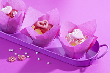 Tablett mit drei Muffins in rosa Verpackungen und Zuckerperlen auf rosa Grund - CSF020979