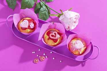 Tablett mit drei Muffins in rosa Verpackungen, Rosenblüten und Zuckerperlen auf rosa Grund - CSF020980