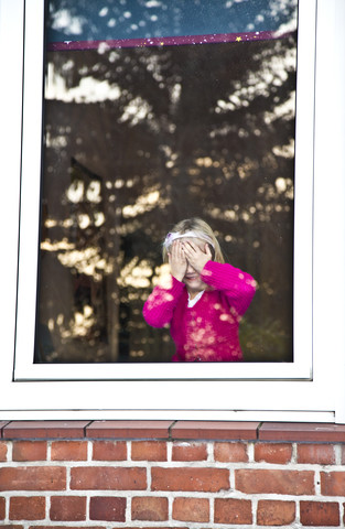 Little girl standing at window covering her eyes stock photo