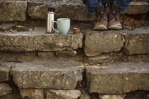 Feet of young man sitting on steps with cup of coffee and thermos flask having a break stock photo