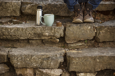 Füße eines jungen Mannes, der mit einer Tasse Kaffee und einer Thermoskanne auf einer Treppe sitzt und eine Pause macht, lizenzfreies Stockfoto