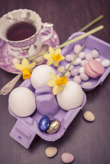 Gedeckter Tisch mit Tasse Tee, Narzissen (Narcissus pseudonarcissus), Eierkarton mit Macaron, drei Eiern und Süßigkeiten auf Holztisch - VTF000167