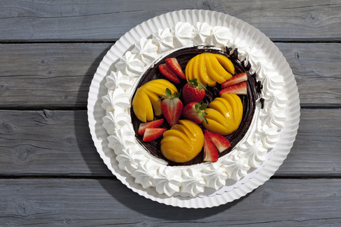 Meringue gebackene Gebäckhülle mit Schokoladenglasur garniert mit Erdbeeren und Pfirsichen auf Pappteller und grauem Holz, lizenzfreies Stockfoto