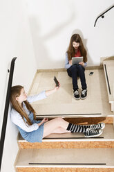 Zwei Mädchen im Teenageralter sitzen auf einer Treppe und benutzen ein Smartphone und ein digitales Tablet - MAEF008279
