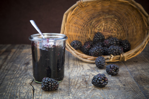 Korb mit Brombeeren (Rubus sectio Rubus) und Einmachglas mit Brombeergelee auf Holztisch, lizenzfreies Stockfoto