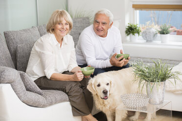 Älteres Paar sitzt nebeneinander auf dem Sofa im Wohnzimmer, Hund im Vordergrund - WESTF019234