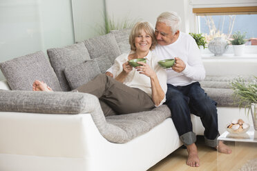 Senior couple with tea cups side by side on sofa in living room - WESTF019236