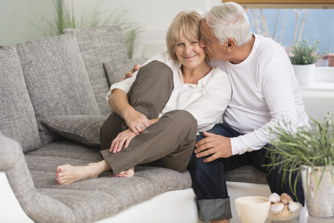 Senior küsst seine Frau auf dem Sofa im Wohnzimmer, lizenzfreies Stockfoto