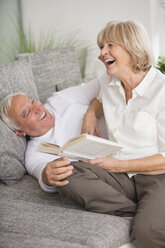 Laughing senior couple with book on sofa in living room - WESTF019248