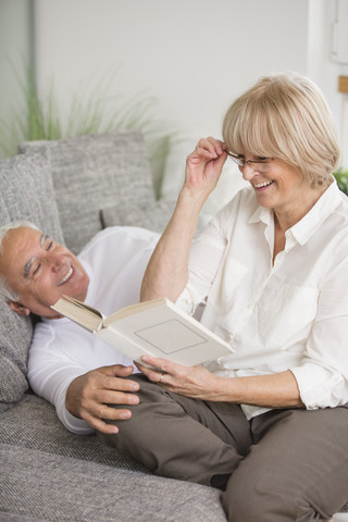 Lächelndes Seniorenpaar mit Buch auf Sofa im Wohnzimmer, lizenzfreies Stockfoto