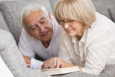 Senior couple with book lying side by side on sofa - WESTF019251