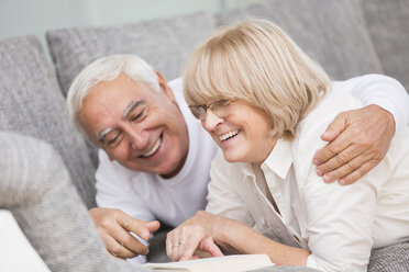 Laughing senior couple with book lying side by side on sofa - WESTF019253