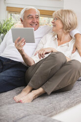 Älteres Paar mit digitalem Tablet nebeneinander auf dem Sofa im Wohnzimmer - WESTF019256