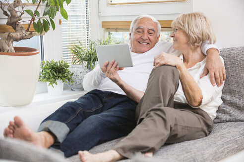 Älteres Paar mit digitalem Tablet nebeneinander auf dem Sofa im Wohnzimmer - WESTF019258