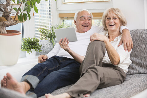 Älteres Paar mit digitalem Tablet nebeneinander auf dem Sofa im Wohnzimmer - WESTF019259