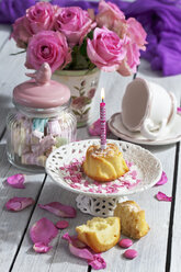 Muffin mit angezündeter Geburtstagskerze, Glas mit Marshmallows, Tasse und rosa Rosen auf dem Tisch - CSF021063