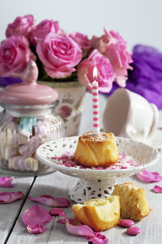 Muffin mit angezündeter Geburtstagskerze, Glas mit Marshmallows, Tasse und rosa Rosen auf dem Tisch, lizenzfreies Stockfoto