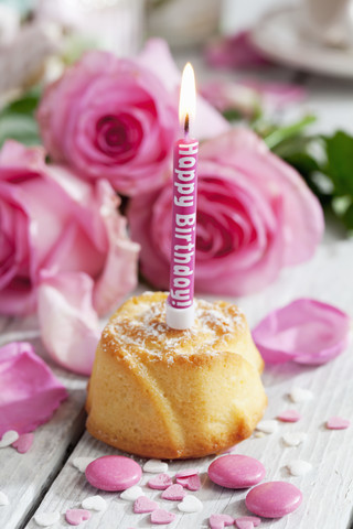 Muffin mit angezündeter Geburtstagskerze, Backdekor und rosa Rosen auf dem Tisch, lizenzfreies Stockfoto