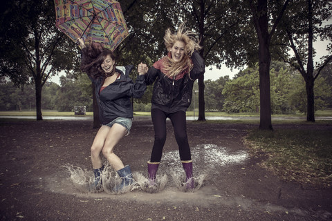 Zwei verspielte junge Frauen mit Regenschirm springen in eine Pfütze, lizenzfreies Stockfoto