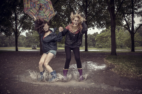 Zwei verspielte junge Frauen mit Regenschirm springen in eine Pfütze - GCF000004