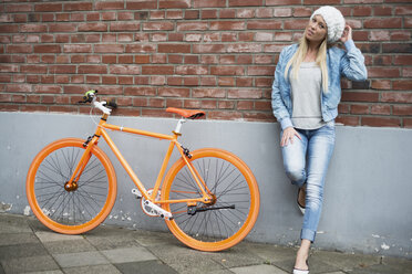 Modische junge Frau lehnt an einer Backsteinmauer neben einem orangefarbenen Fahrrad - LFOF000166