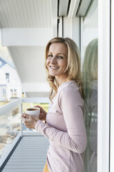 Smiling woman standing on balcony of her house with a cup of coffee - MFF000965