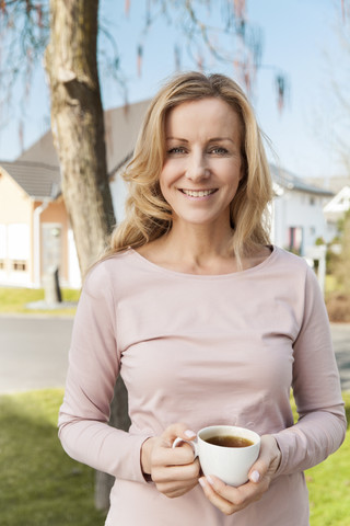 Lächelnde Frau mit einer Tasse Kaffee vor einem Wohngebiet stehend, lizenzfreies Stockfoto