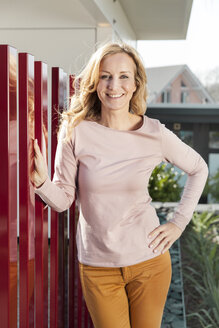 Lächelnde Frau steht am roten Zaun eines Wohnhauses - MFF000980