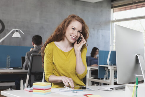 Porträt einer lächelnden Frau, die mit ihrem Smartphone an ihrem Arbeitsplatz im Großraumbüro telefoniert - RBF001682