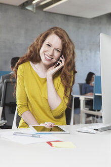 Porträt einer lächelnden Frau, die mit ihrem Smartphone an ihrem Arbeitsplatz im Großraumbüro telefoniert - RBF001620