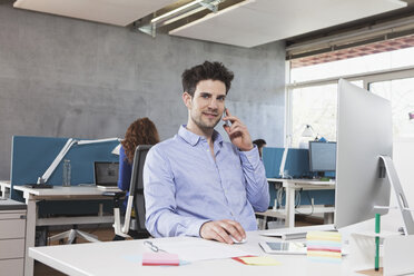 Porträt eines Mannes, der mit seinem Smartphone an seinem Arbeitsplatz im Büro telefoniert - RBF001626