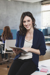 Porträt einer lächelnden Frau mit digitalem Tablet, die an ihrem Arbeitsplatz in einem Großraumbüro sitzt - RBF001643