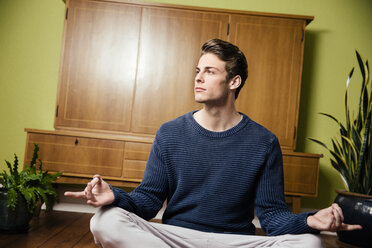 Germany, Bonn, Young man in a yoga pose, sitting on the wooden floor of his home, looking away - MFF001011