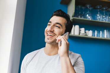 Porträt eines lachenden jungen Mannes, der in seiner Küche mit seinem Smartphone telefoniert - MFF001022