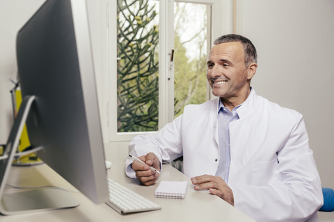 Lächelnder Arzt sitzt am Schreibtisch mit Computer, lizenzfreies Stockfoto