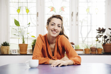 Porträt einer lächelnden jungen Frau, die zu Hause am blauen Tisch sitzt - MFF001047