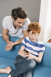 Father looking over shoulder of his son who is using a smart phone at home - MFF001077